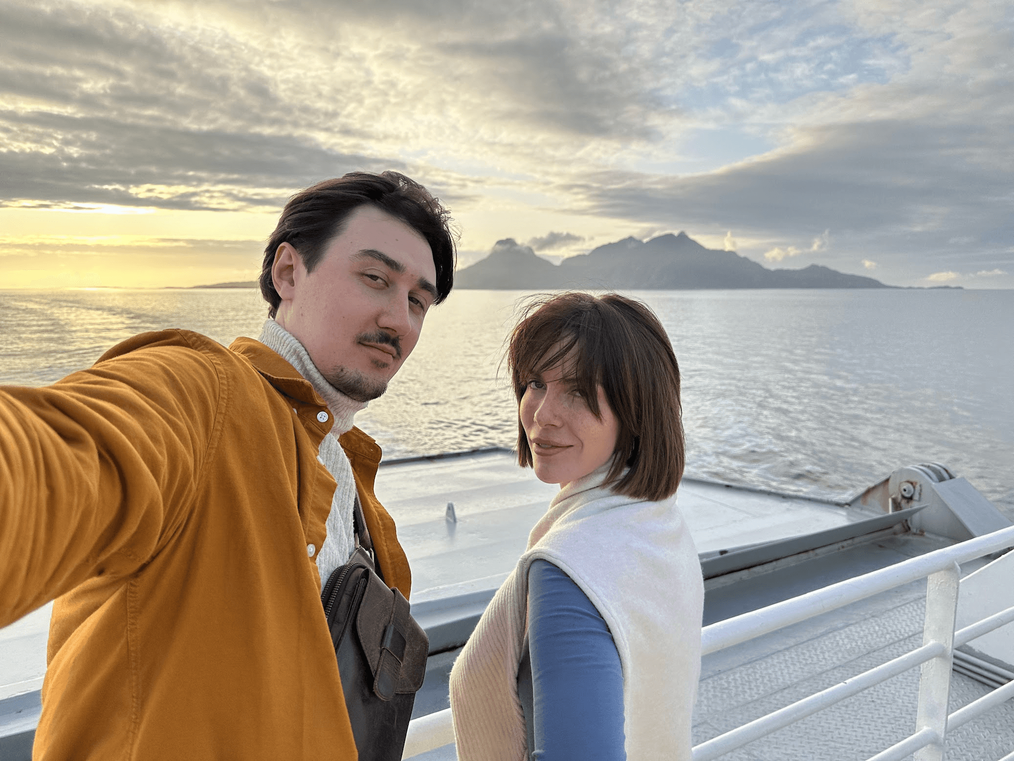 A couple takes a selfie on a boat with a scenic sunset and mountains in the background.