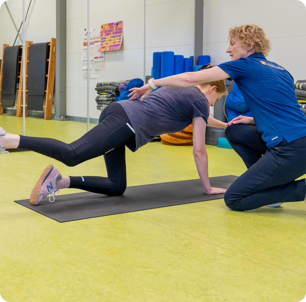 A woman in a gym assists another woman performing a balancing exercise on a yoga mat, surrounded by fitness equipment, emphasizing exercise, balance, and physical therapy.