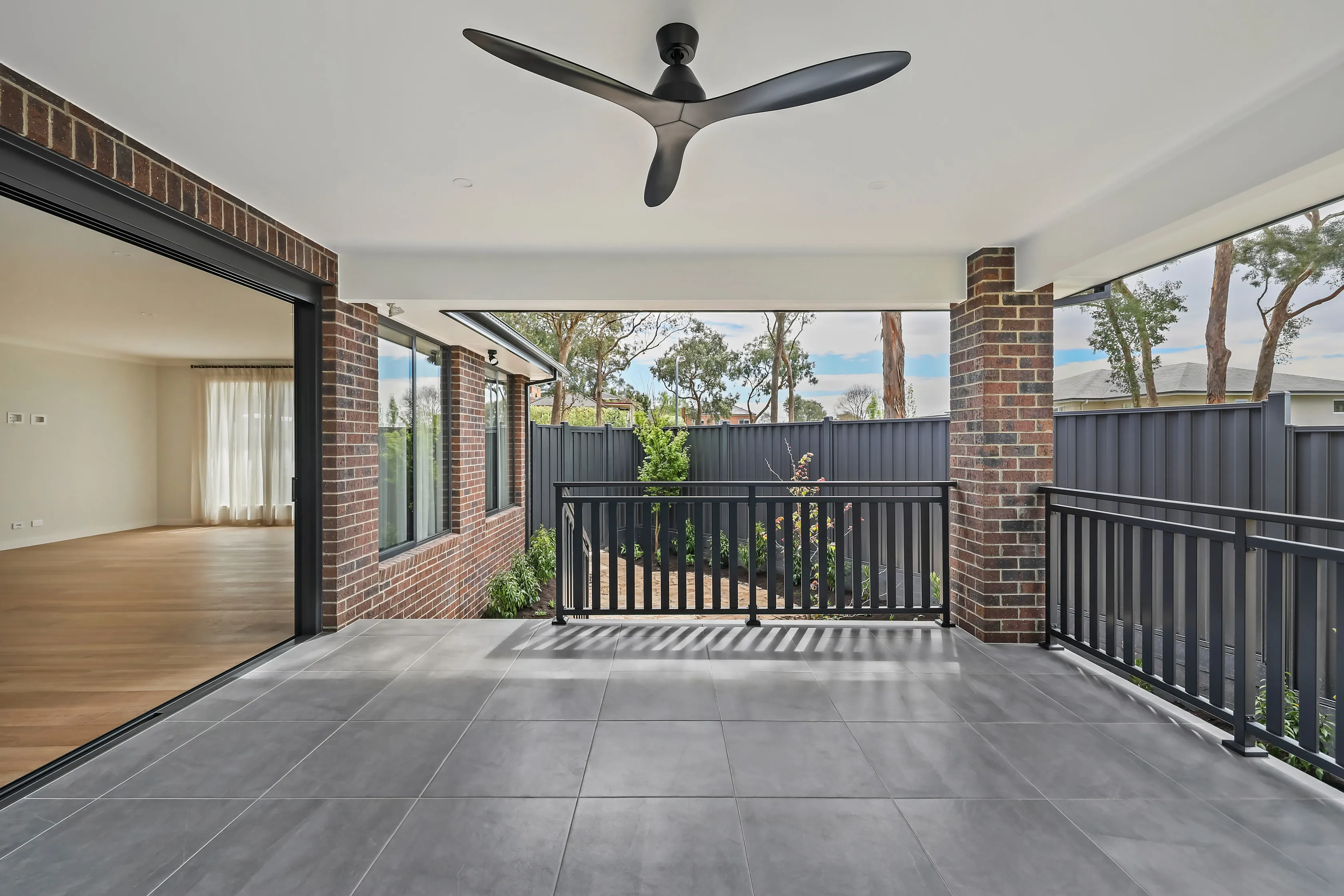 Covered alfresco area with grey tiled floor, a black ceiling fan, brick piers, a black balustrade and a black fence, with sliding doors back to the living room and a bush outlook