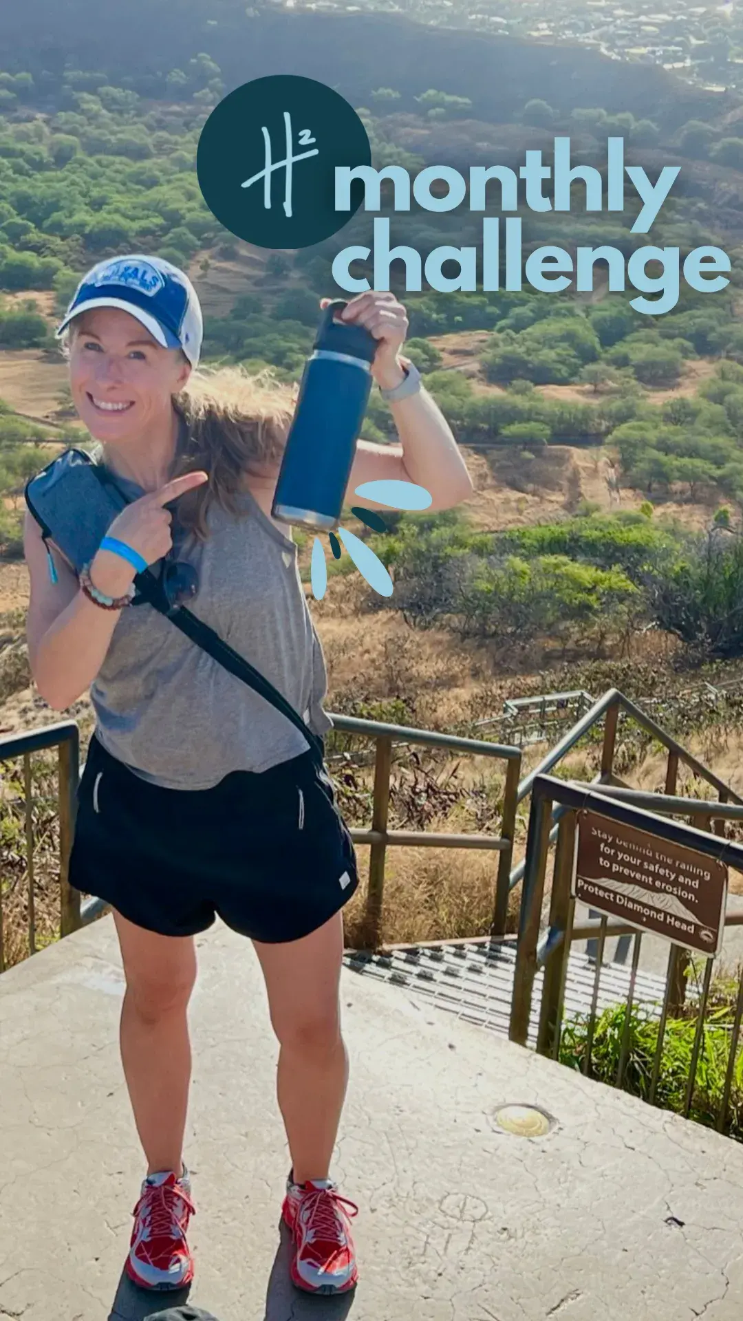 Heather Hausman in athletic wear stands on a hiking trail with lush green foliage and a clear blue sky in the background, holding up a blue water bottle as part of a "monthly challenge" promotion.
