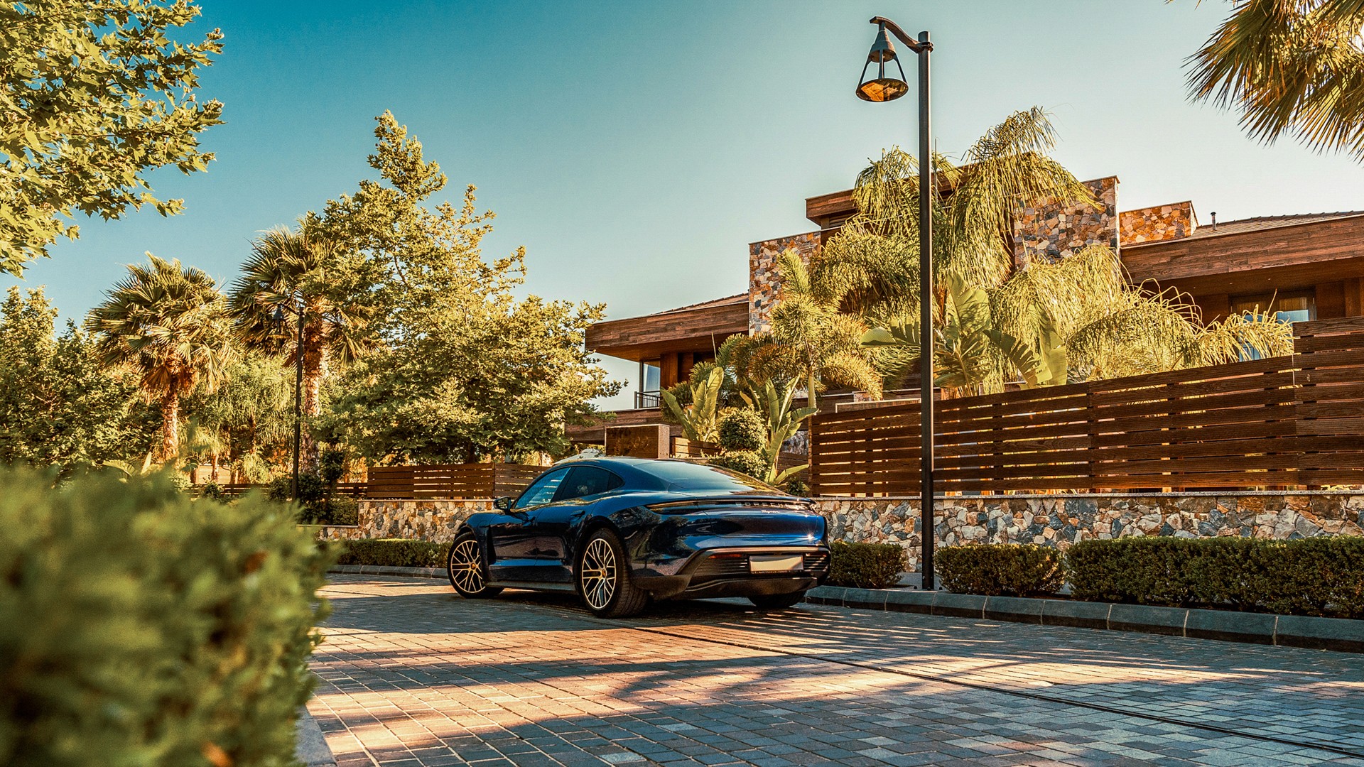 Dark blue Porsche Taycan parked on a block-paved driveway beside a modern stone-and-timber villa, palm trees and a coach-style lamp post around it in warm afternoon light.
