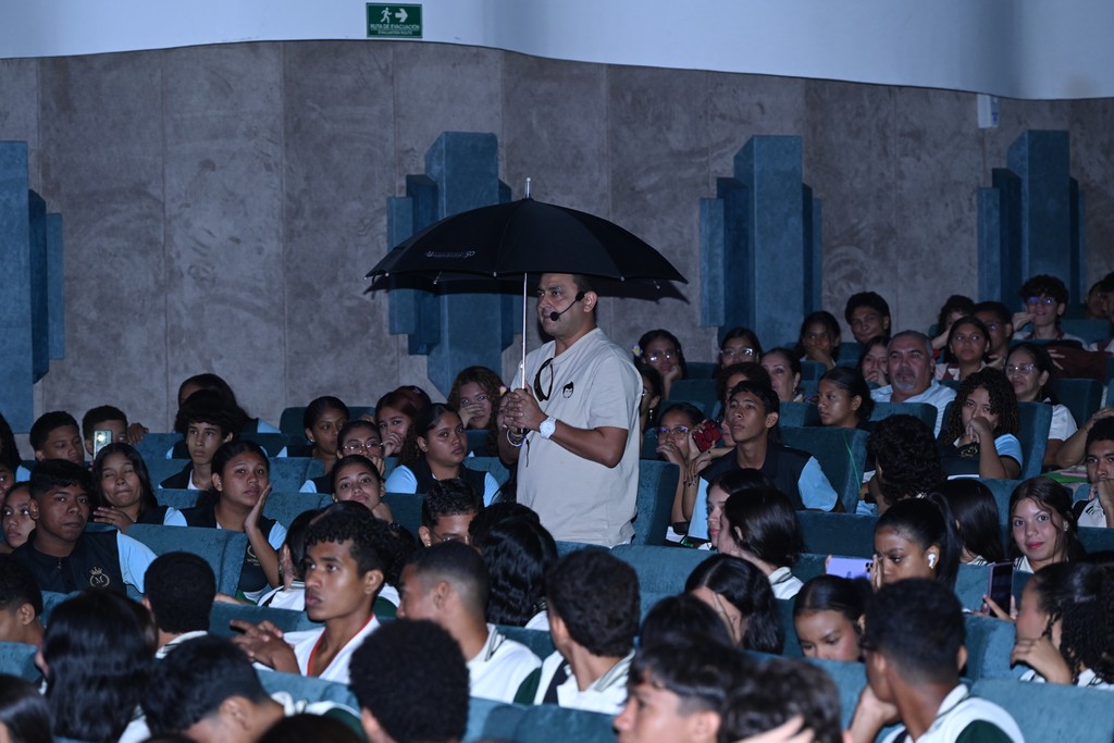 Jorge Baena caminando entre estudiantes con un paraguas negro abierto, mientras da una charla interactiva en un auditorio.