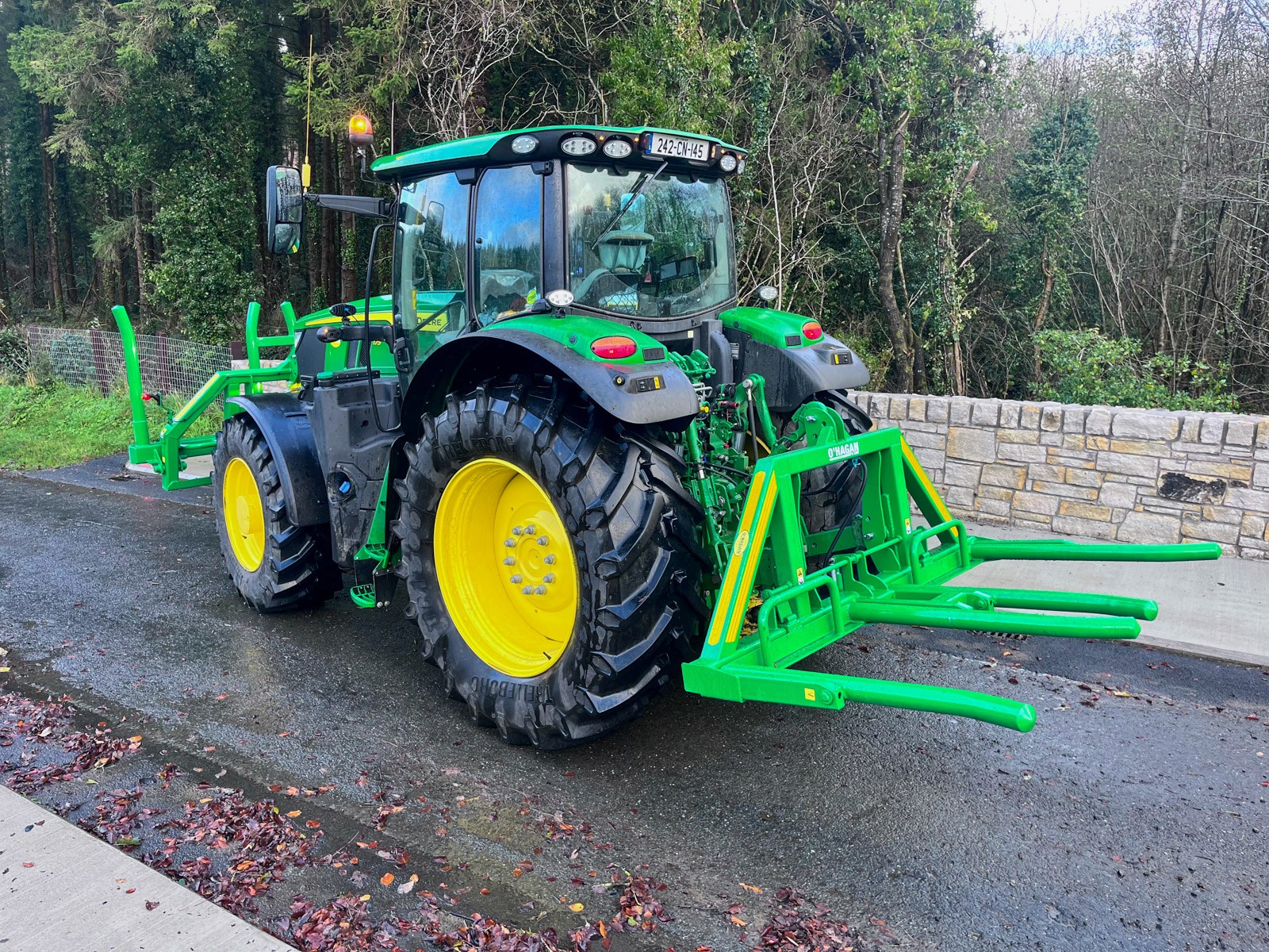 John Deere Green Tractor with black and yellow wheels on a country road with a green hydraulic bale handler attached beside an old wall with big trees in the background