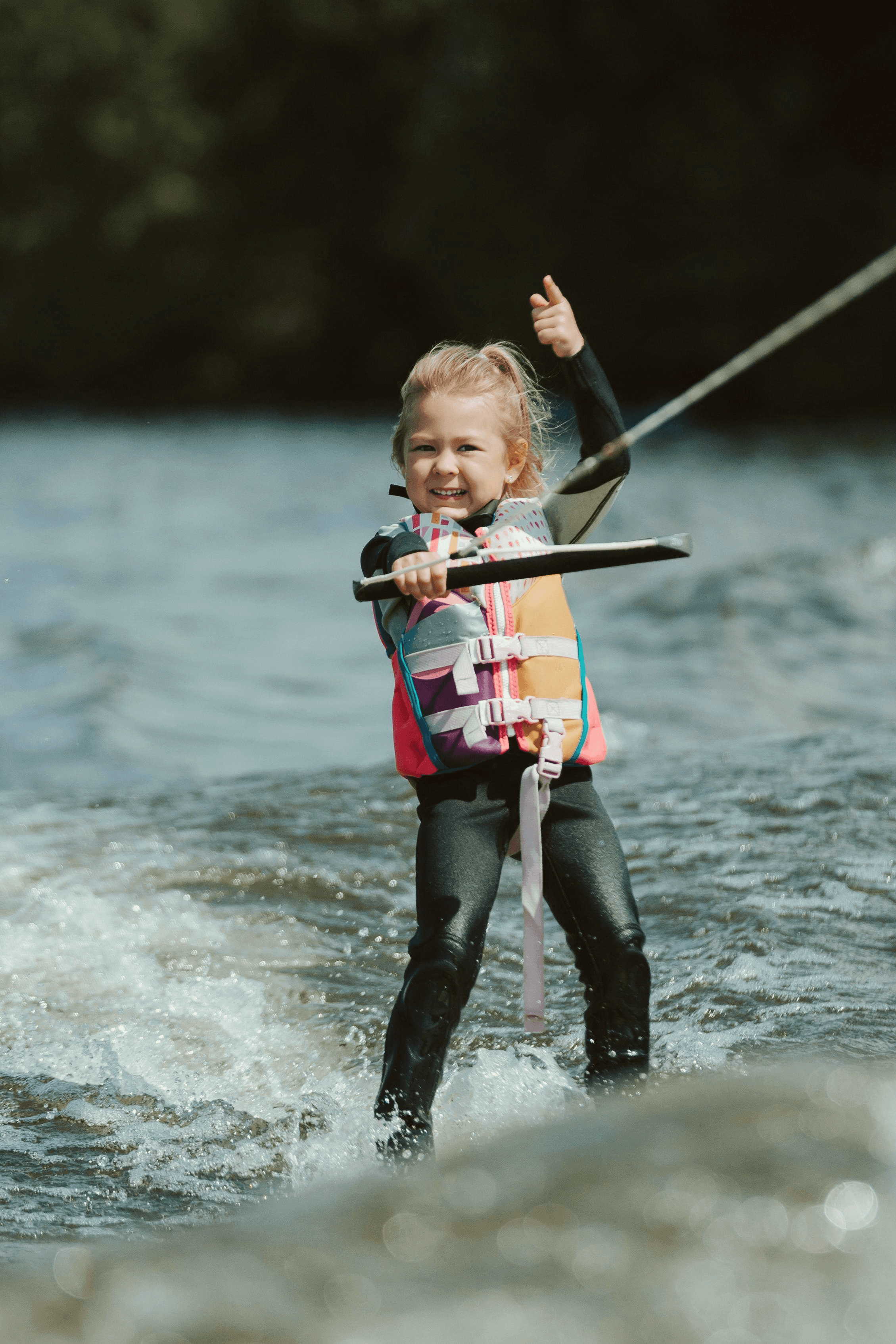 A young child wearing a colorful life jacket and black wetsuit is confidently water skiing on a lake, giving a thumbs-up while holding onto the tow rope, with splashes of water around them and a blurred natural background.