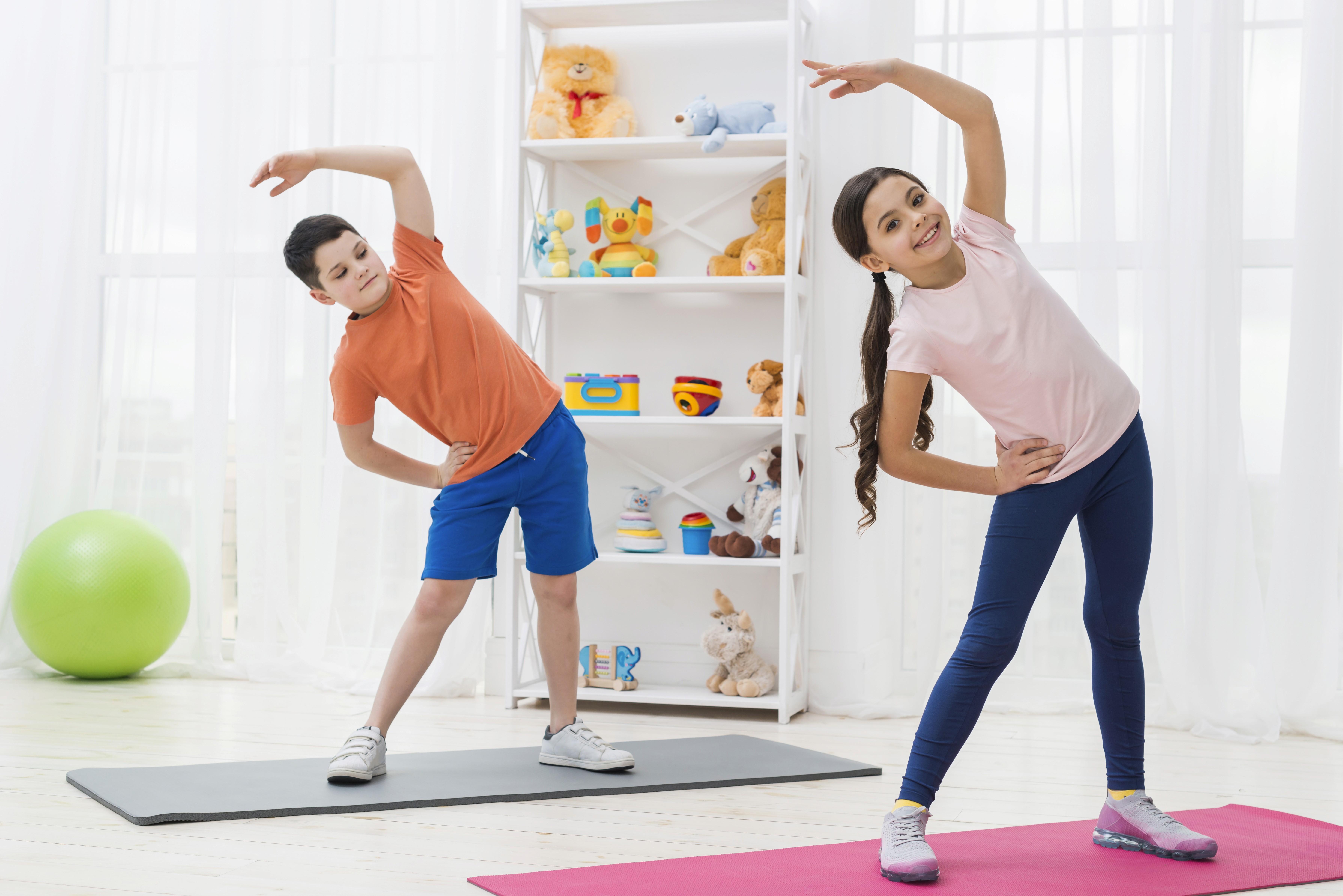 Two children are stretching on yoga mats in a bright room, smiling. A white shelf with colorful toys is in the background, creating a playful atmosphere.