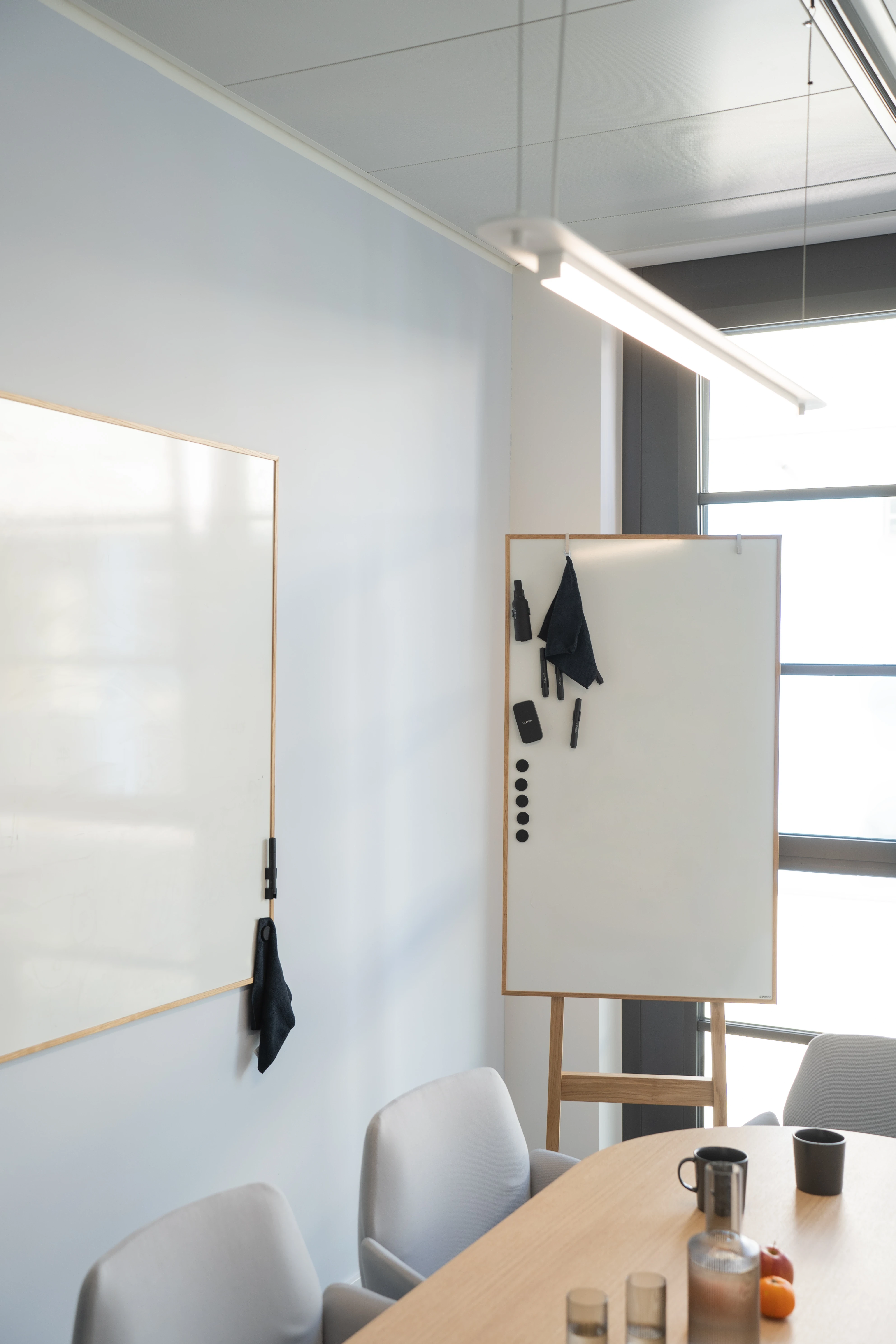 Meeting room corner at AI START-UP MUNICH with a wall-mounted whiteboard and a mobile whiteboard easel in oak, grey upholstered chairs and a wooden table with carafe and fruit.