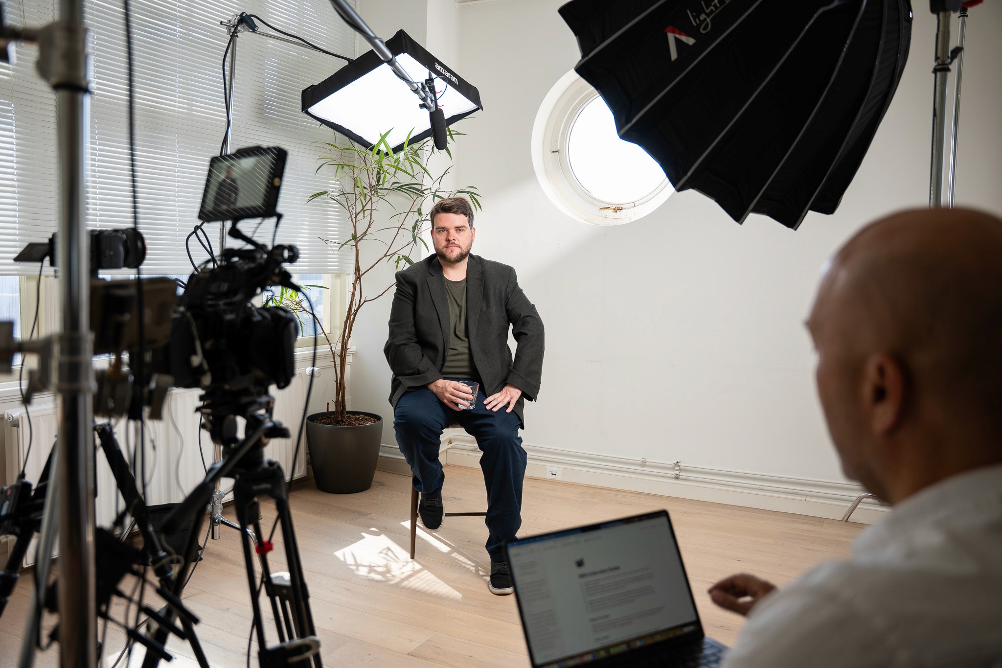 Wide shot of a man in a dark blazer seated on a stool during a video shoot, with two cameras, softbox lighting rigs, and a boom microphone visible, and an interviewer with a laptop in the foreground.