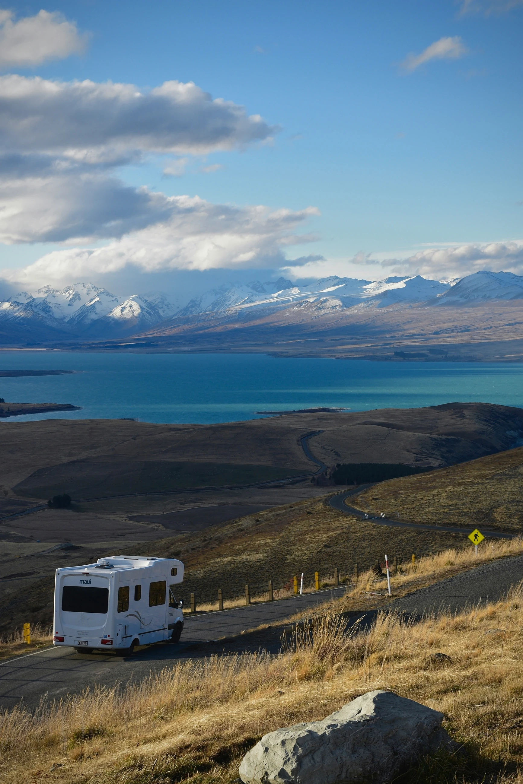 A white motorhome driving on a scenic mountain road overlooking a turquoise lake and snow-capped peaks.