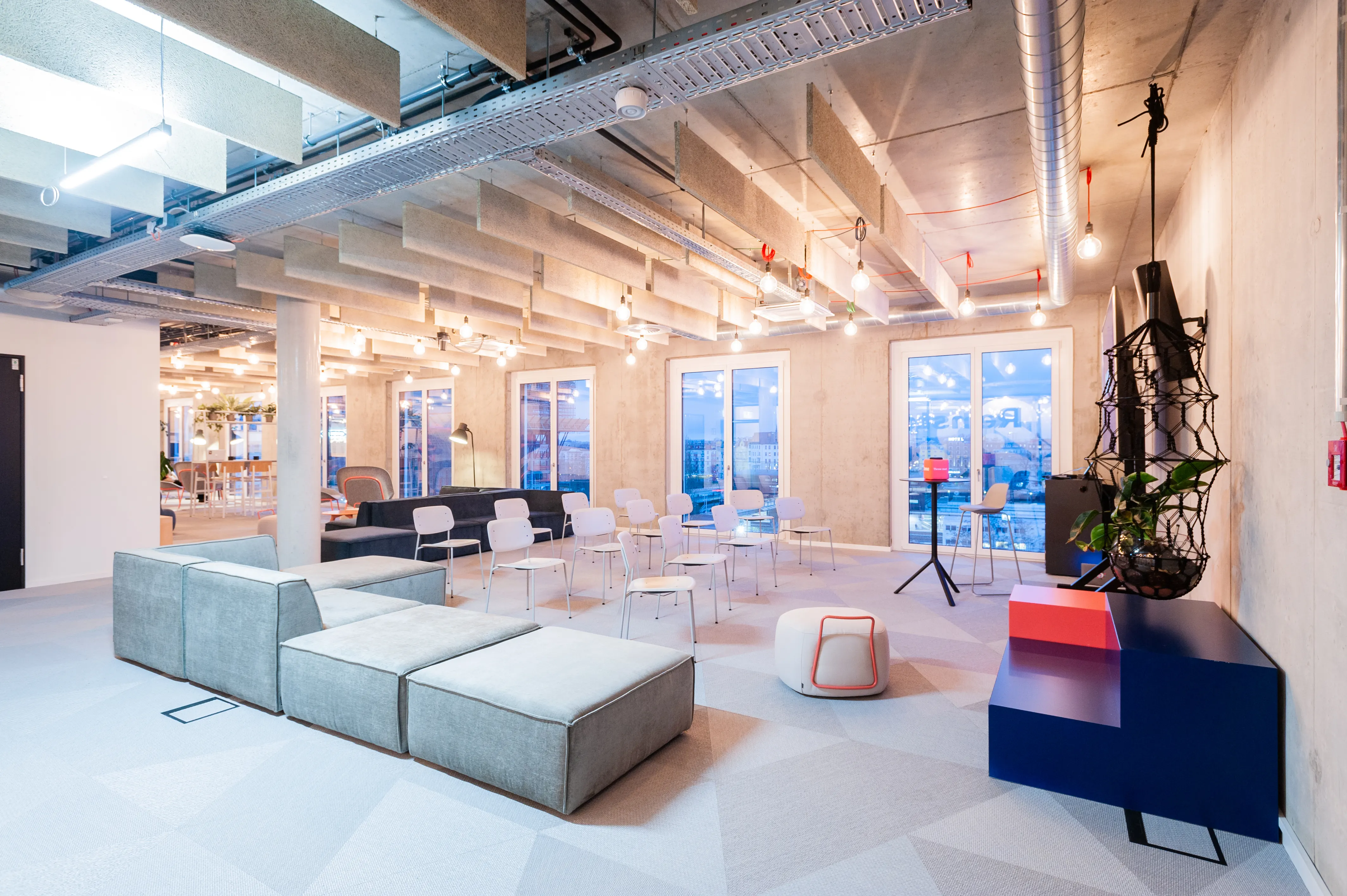 Event lounge at OMIO with light grey modular sofas, white chairs arranged loosely on a pale geometric carpet, a black net sculpture with plants and a navy-and-red sideboard.
