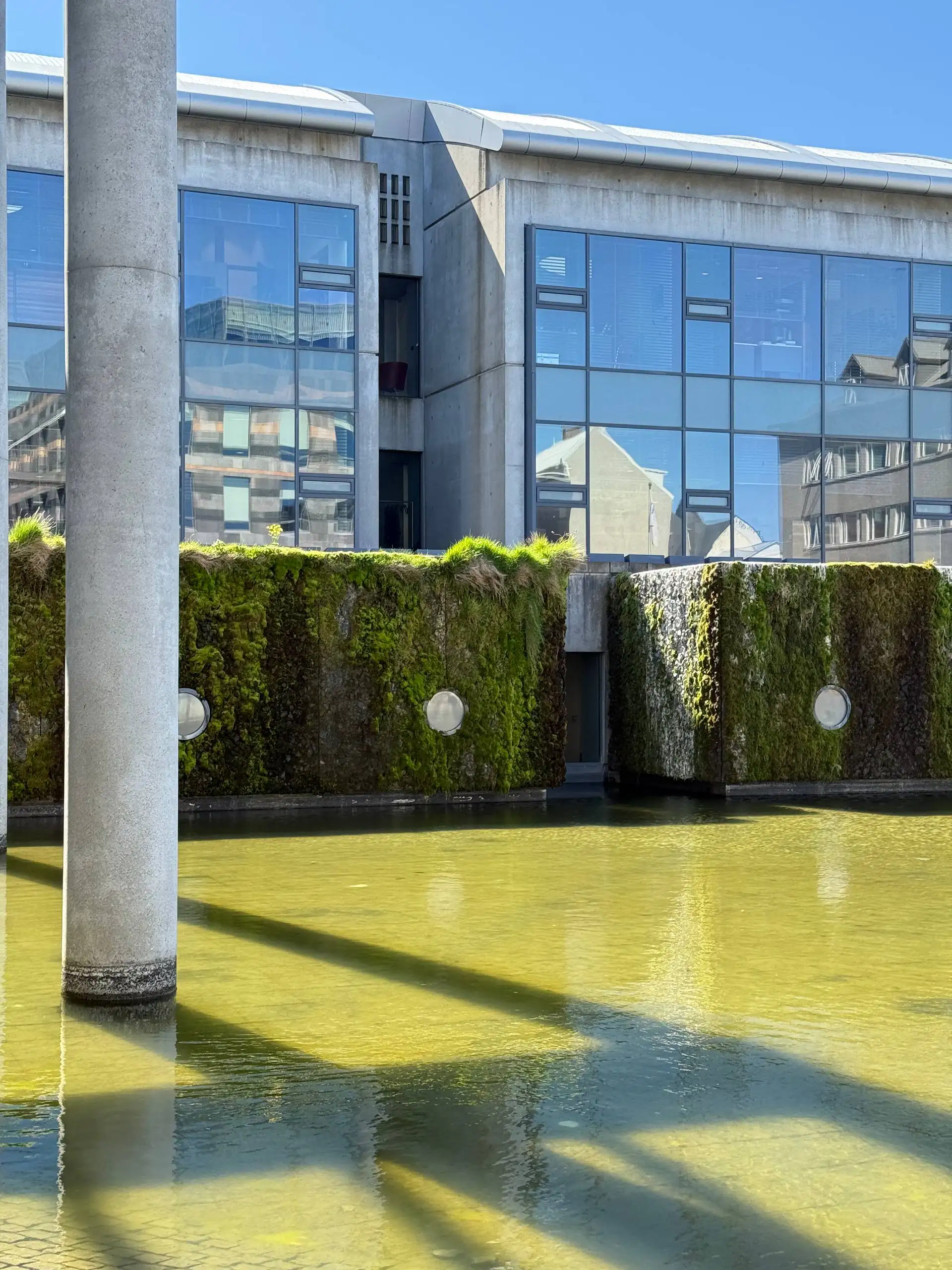 Reykjavík City Hall’s concrete and glass façade rising above the still green water of The Pond, with moss-covered walls at the water’s edge