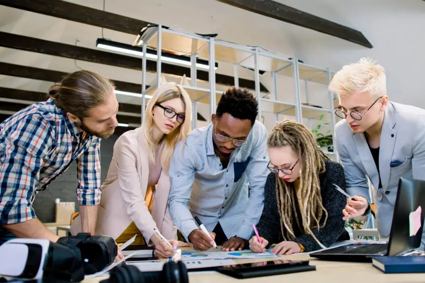 A group of five people is gathered around a desk, examining and discussing a document. They are actively taking notes and collaborating, suggesting a work meeting or a brainstorming session.