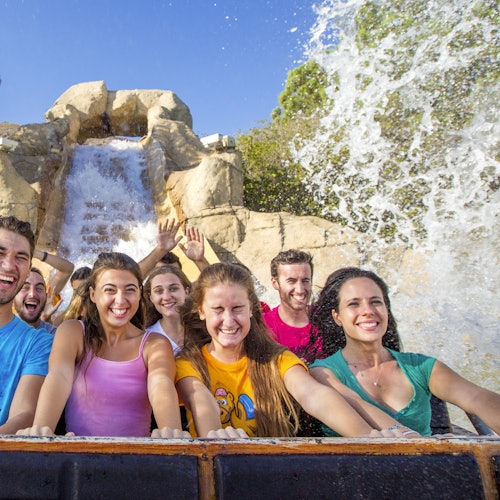 Grupo de personas sonrientes montando un canal de troncos con agua salpicando contra un fondo rocoso bajo un cielo azul claro.