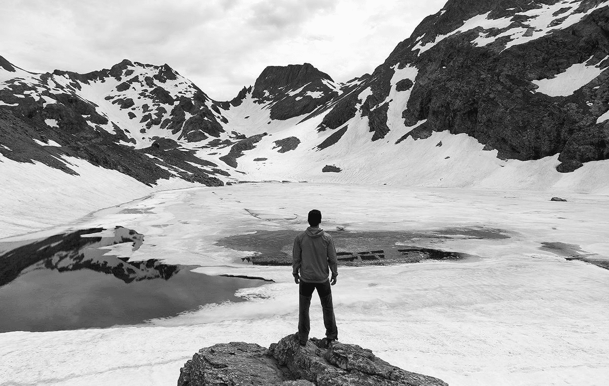 A person standing before a frozen mountain lake, surrounded by rugged snow-covered peaks — a scene of solitude, awe, and natural strength.