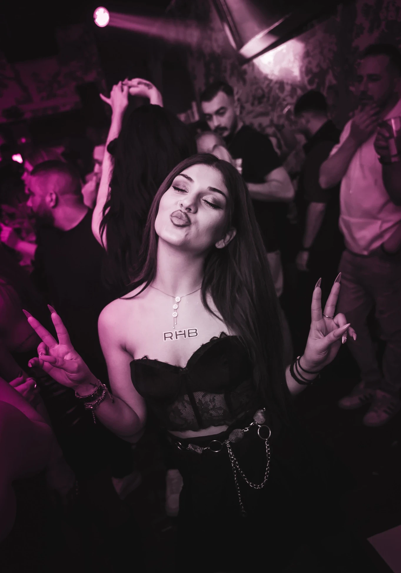 A woman in a black strapless top and an "RHB" necklace looks at the camera, making a kissy face and flashing peace signs with both hands in a crowded, pink-lit club.