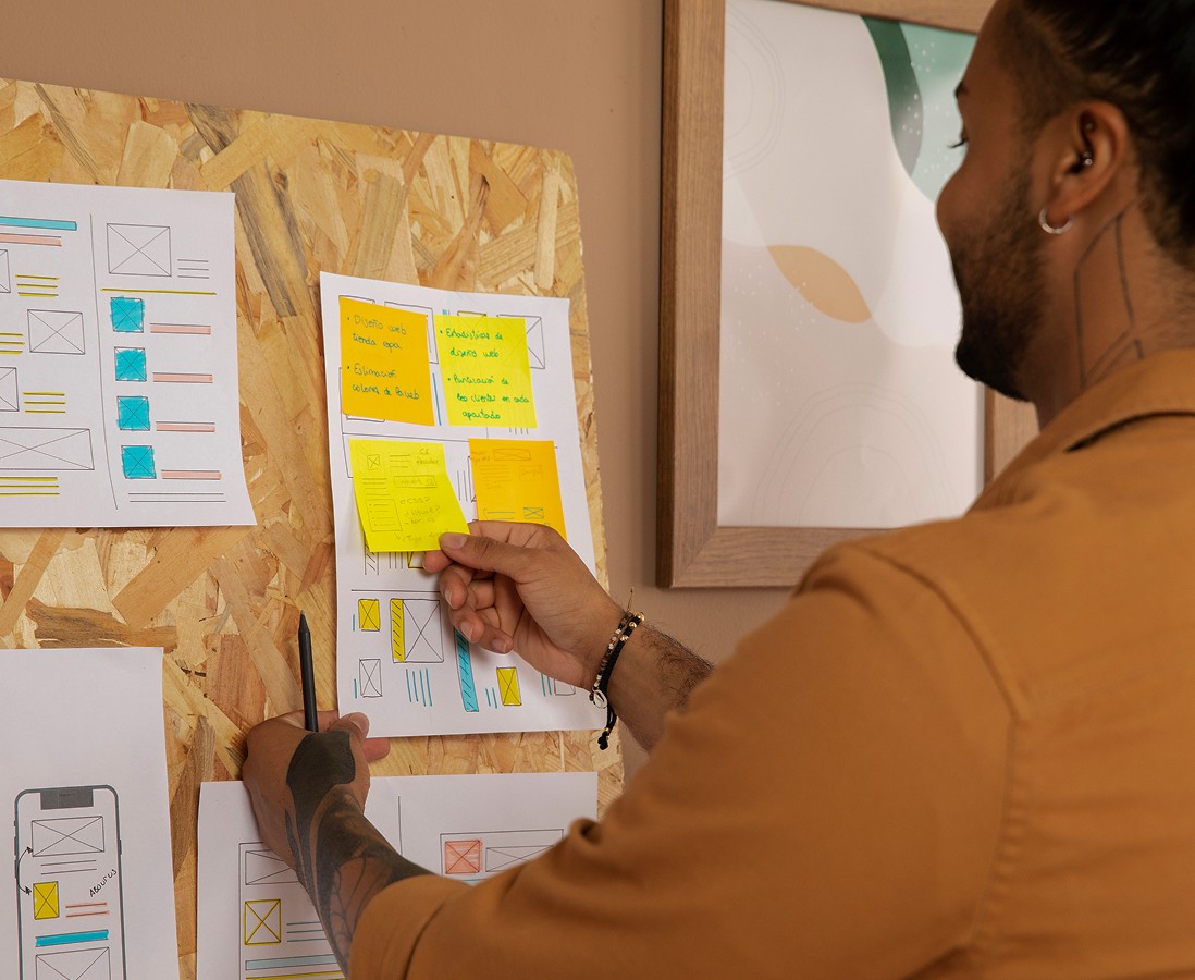 A person reviews colorful sticky notes and sketches of a mobile app design on a corkboard, focusing on wireframes and layout drafts.