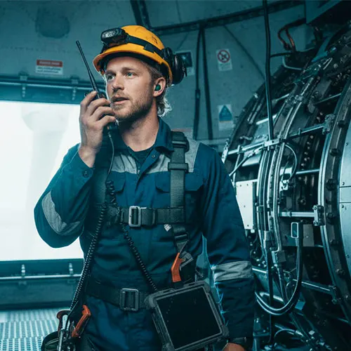 Field technician wearing a safety harness and helmet communicating via two-way radio inside a turbine or mechanical room.