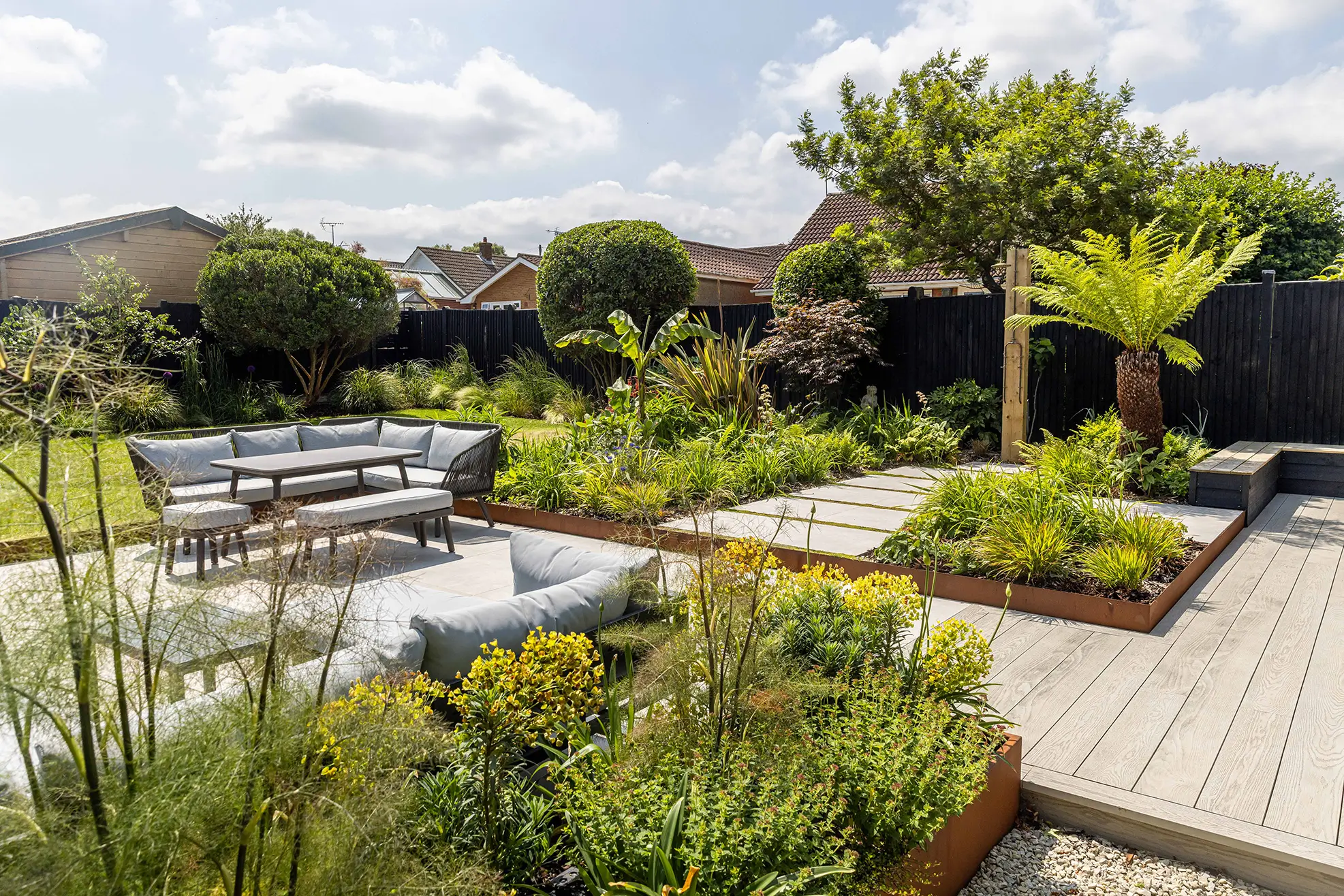 Lush garden with varied plants and pathways, featuring a seating area under a blue sky with fluffy clouds.
