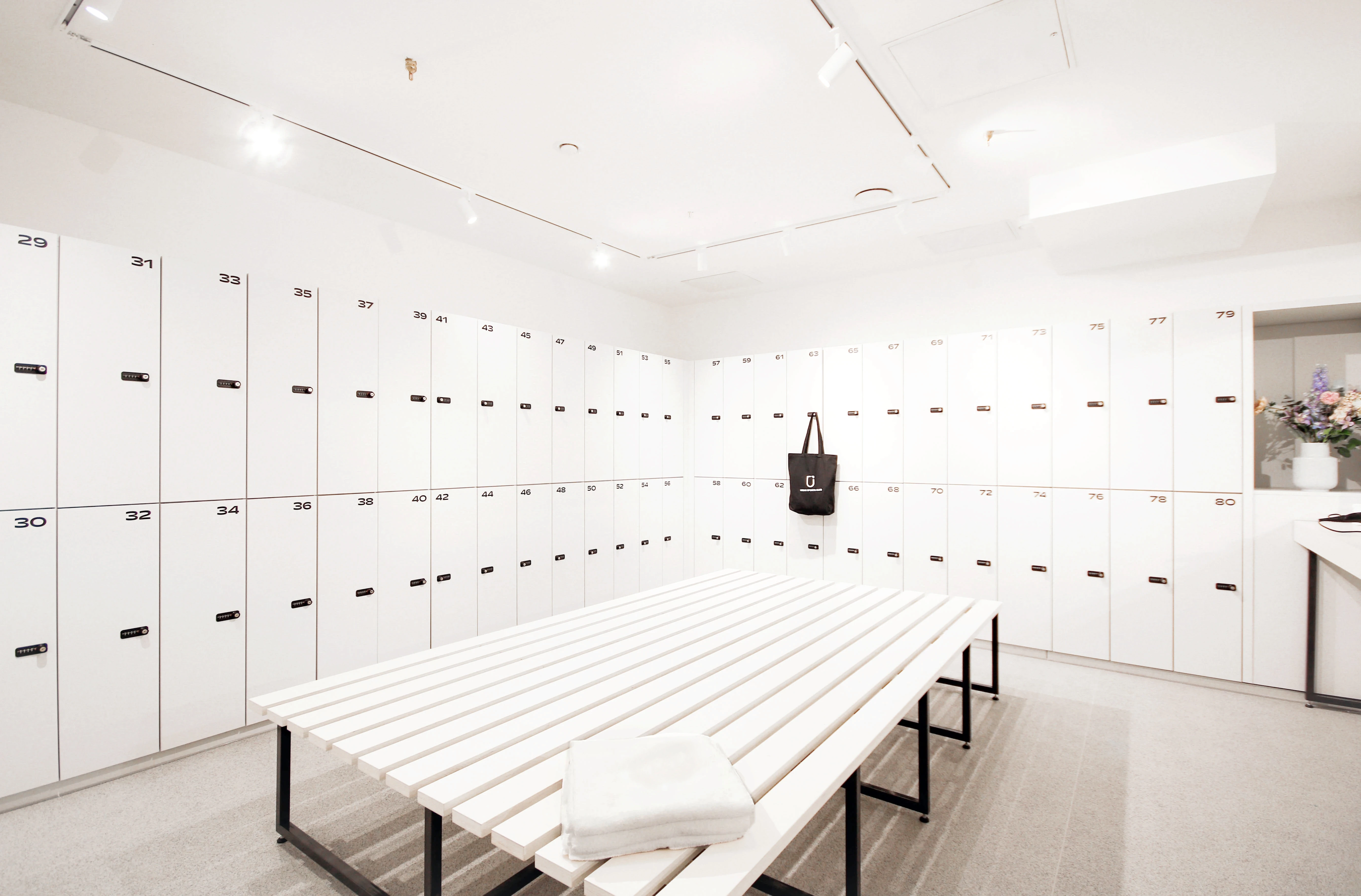 Bright changing room at CYCLA with white numbered lockers along the walls and a long white slatted bench with folded towels in the middle.