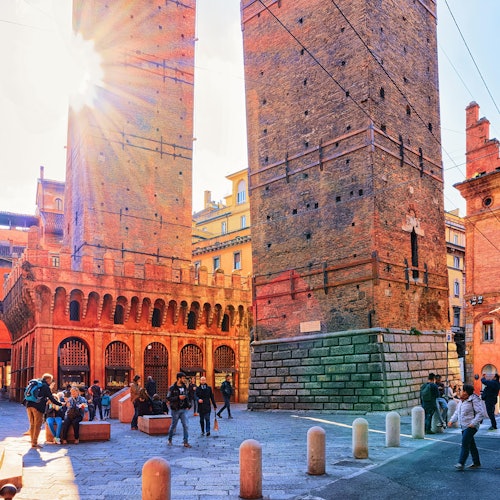 Persone che camminano e si radunano vicino alle storiche torri gemelle di mattoni sotto un cielo soleggiato, con la luce del sole che filtra attraverso una delle torri.