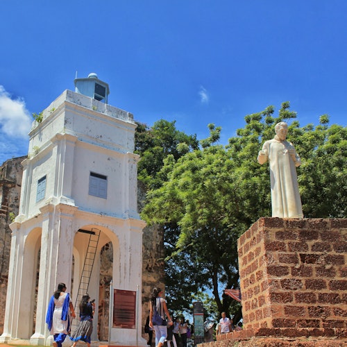 People walking near a tall white bell tower and a statue on a stone pedestal, with trees and a blue sky in the background.