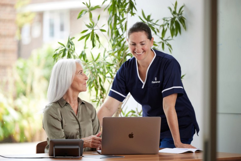 A smiling nurse assisting an elderly woman using a laptop in a bright outdoor setting, representing a positive and supportive working culture in care