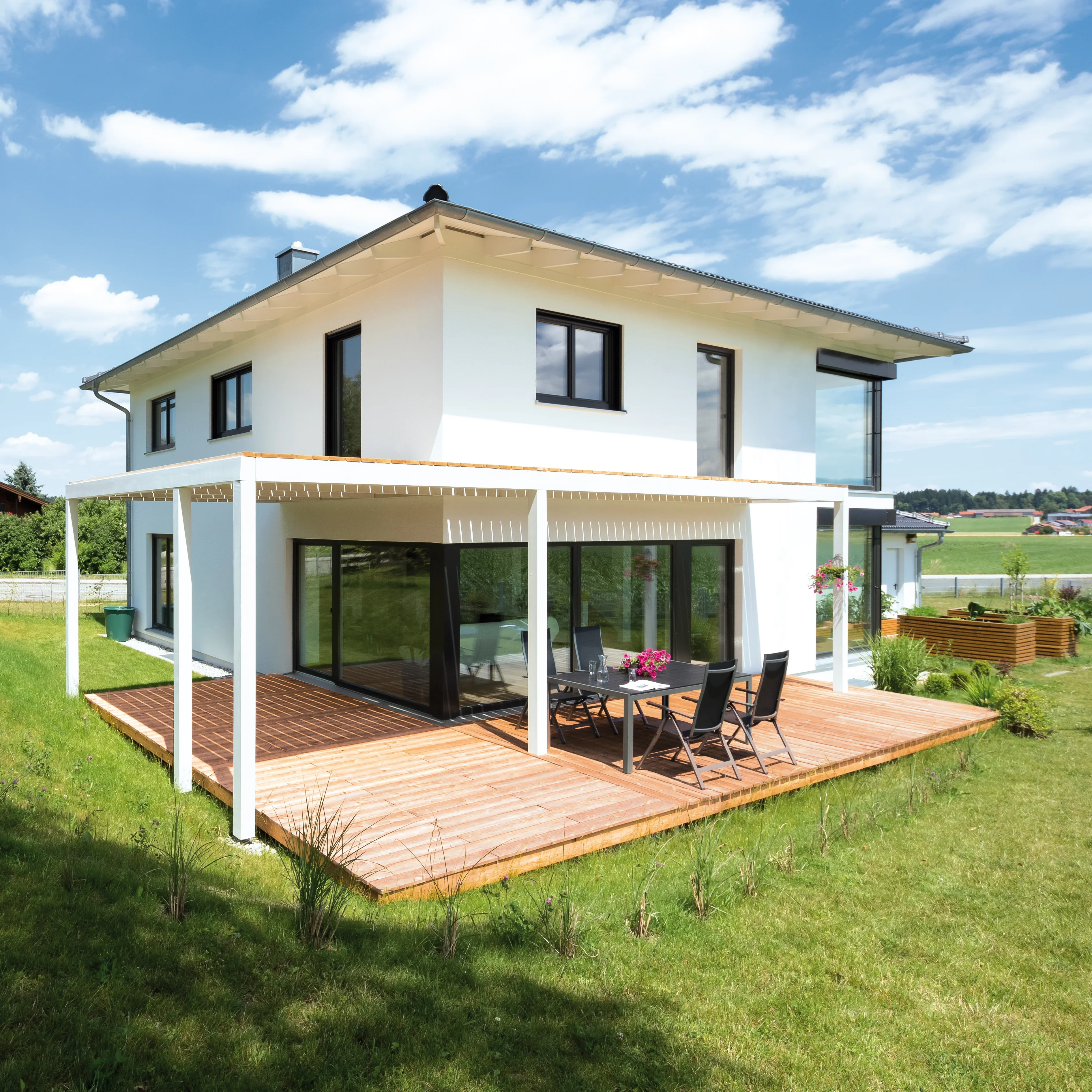 Modernes weißes Haus mit Holzterrasse, Tisch und Stühlen, umgeben von einem gepflegten Garten unter blauem Himmel.