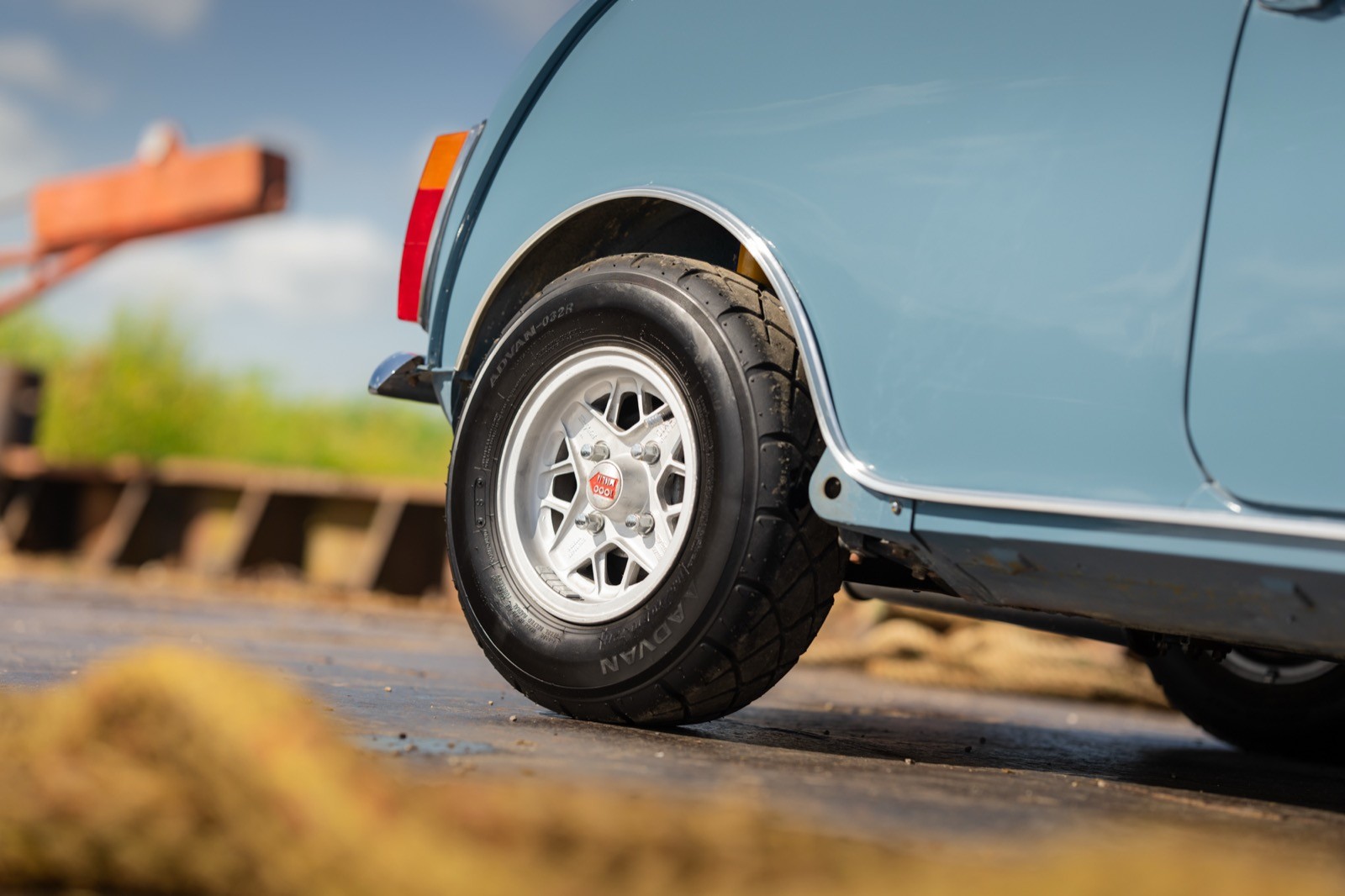Close-up of a blue car's wheel on a road, with a blurred background of greenery and construction equipment.