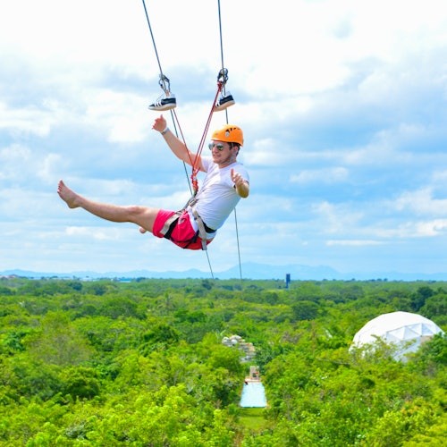 Una persona con casco y gafas de sol está haciendo una tirolesa sobre un bosque verde exuberante, con un cielo nublado de fondo.