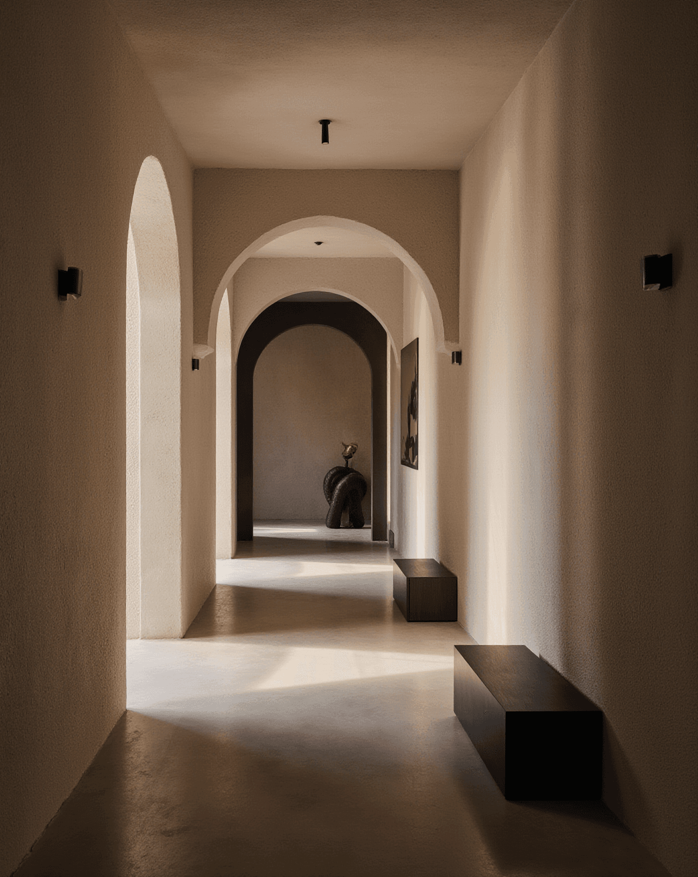 Long hallway with soft light, arched doorways, and dark benches leading to a sculptural figure at the far end.