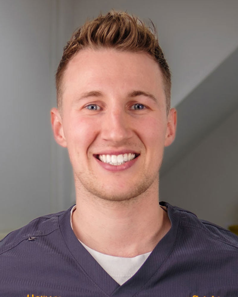 A portrait of Harvey, an Associate Dentist at Cricklade Dental Practice, smiling and wearing a dark grey scrub top with yellow embroidered text that reads "Harvey" on the left and "Cricklade Dental Practice" on the right. He is standing in a dental surgery with a clock and equipment visible in the background.
