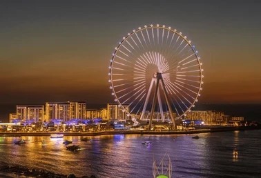 A wide-angle shot of the Ain Dubai Ferris wheel illuminated at sunset/dusk on Bluewaters Island, with city buildings and boats in the water.