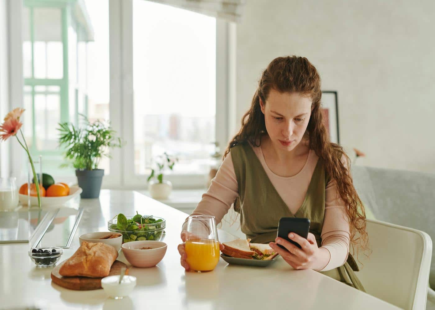 Woman at kitchen table with a sandwich in front of her holding a glass of juice while looking at smartphone