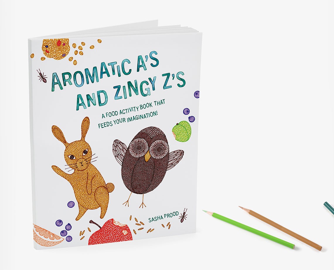The image shows a children's activity book titled "Aromatic A's and Zingy Z's: A Food Activity Book That Feeds Your Imagination!" by Sasha Prood. The book cover features illustrations of a rabbit, an owl, and a small insect, along with a lemon, apple, and blueberries. The title is written in teal text on a white background. The book is open, revealing a white interior, and is set against a white background. Two colored pencils are placed to the right of the book.