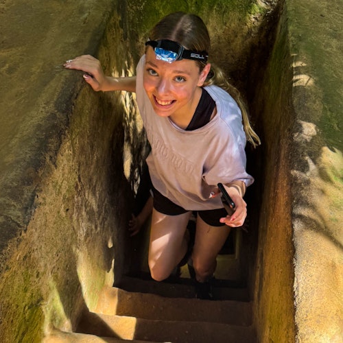 Seorang wanita dengan lampu kepala tersenyum saat menaiki tangga batu yang sempit. Dia memegang ponsel cerdas dan mengenakan kemeja berwarna terang.