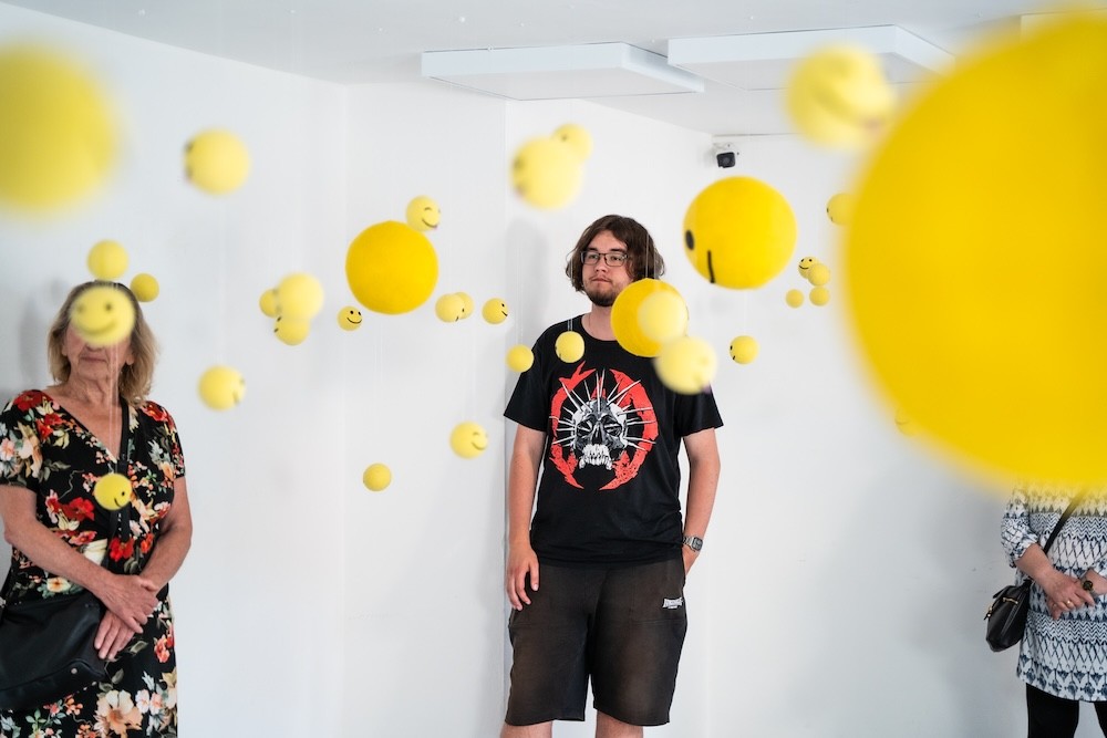 Viaceré žlté smiley objekty visiace v bielom priestore – dielo Niny A. Šoškovej
