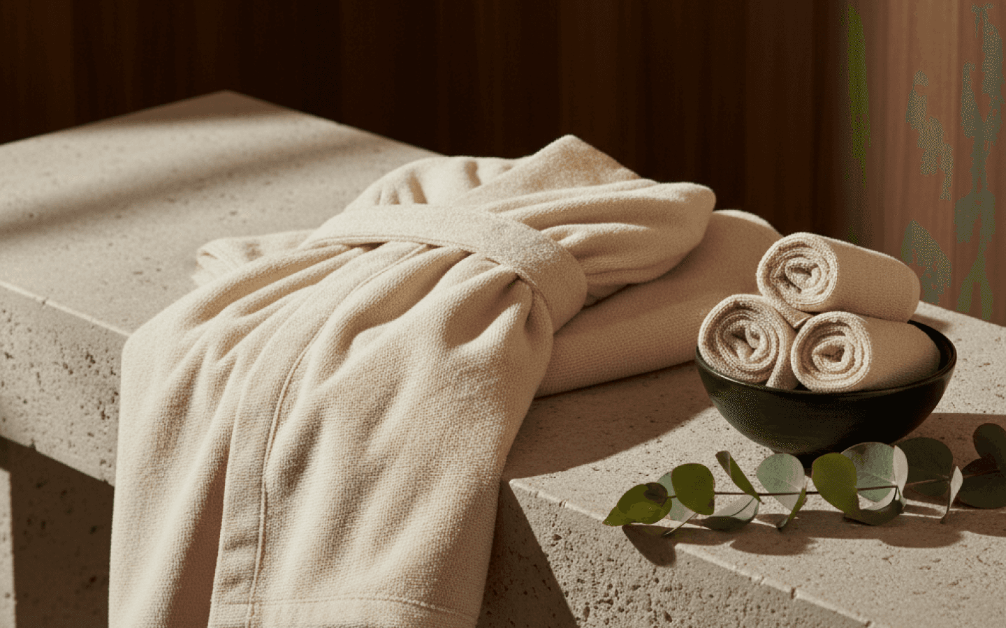 Folded spa wrap and bath sheet on a stone spa bench with rolled hand towels and eucalyptus sprigs