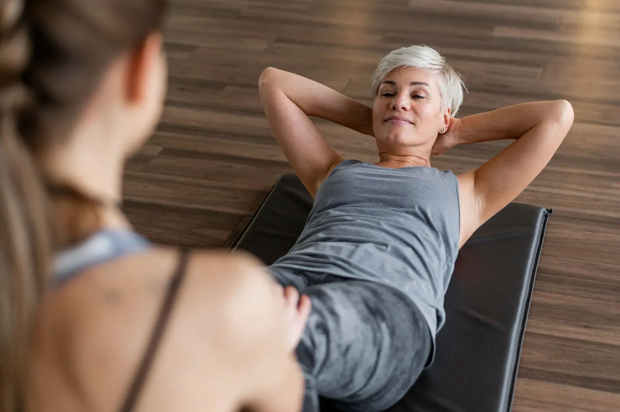 Older woman performing a guided abdominal exercise on a mat with a personal trainer, focusing on strength and core engagement.