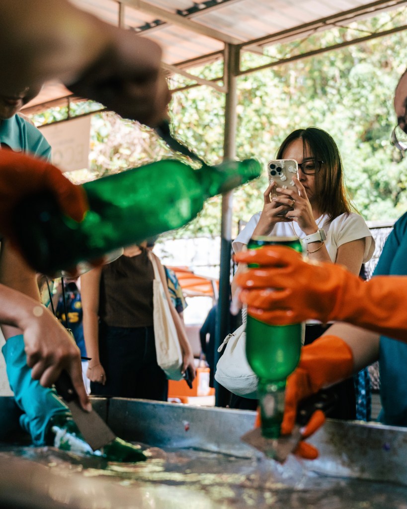 A group of individuals cleaning and preparing green glass bottles for recycling, with one person taking a photo. Bright colors and lively atmosphere reflect a community effort towards sustainability.