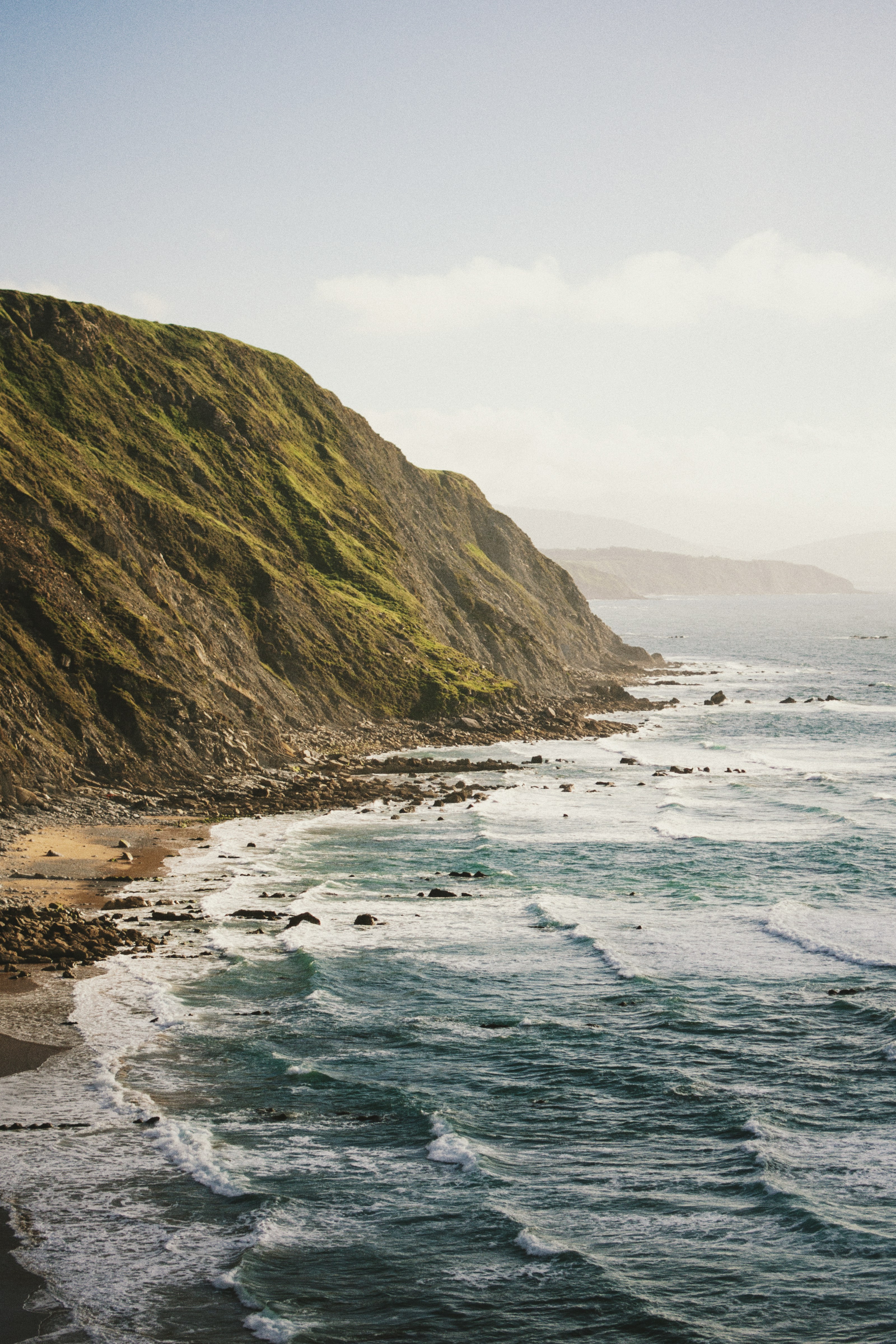 A scenic coastal landscape featuring a rocky shoreline and steep cliffs covered in lush green vegetation, with gentle ocean waves lapping against the shore under a clear, sunny sky.