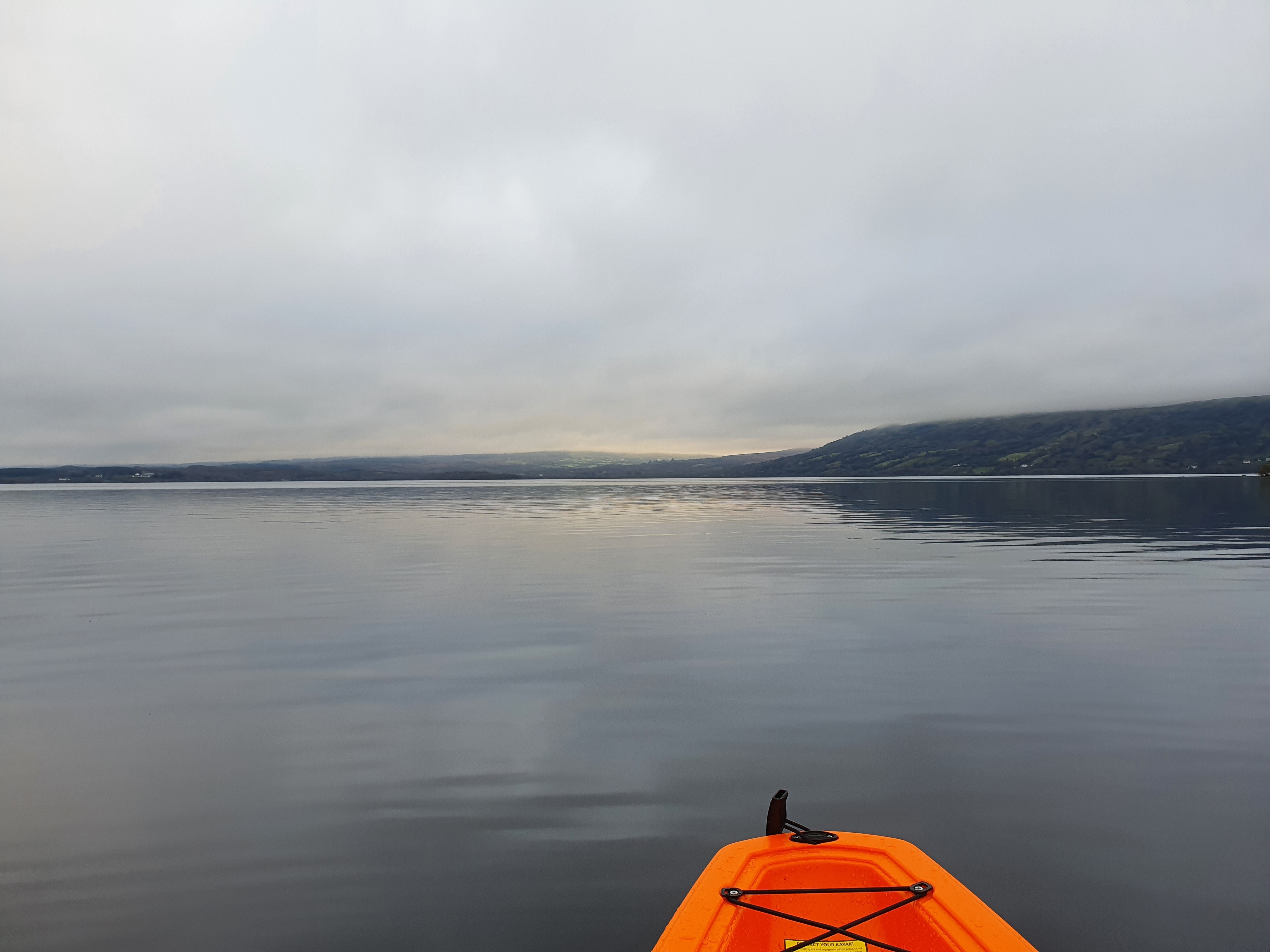 An orange kayak floats on calm waters under a gray sky, with distant hills faintly visible in the background.