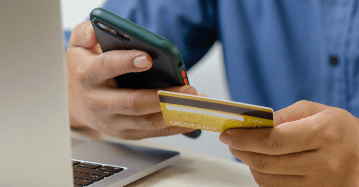 Closeup of a person’s hands holding a credit card and a cell phone in front of a laptop computer.