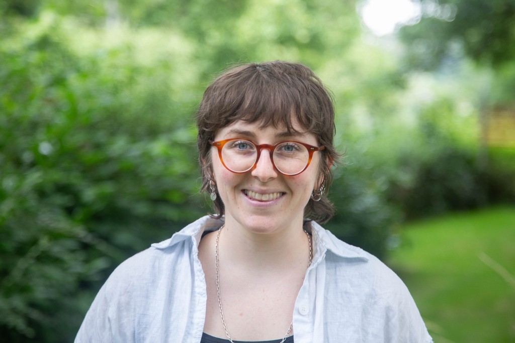 A woman wearing orange glasses smiling infront of a green blurry background of a bush.