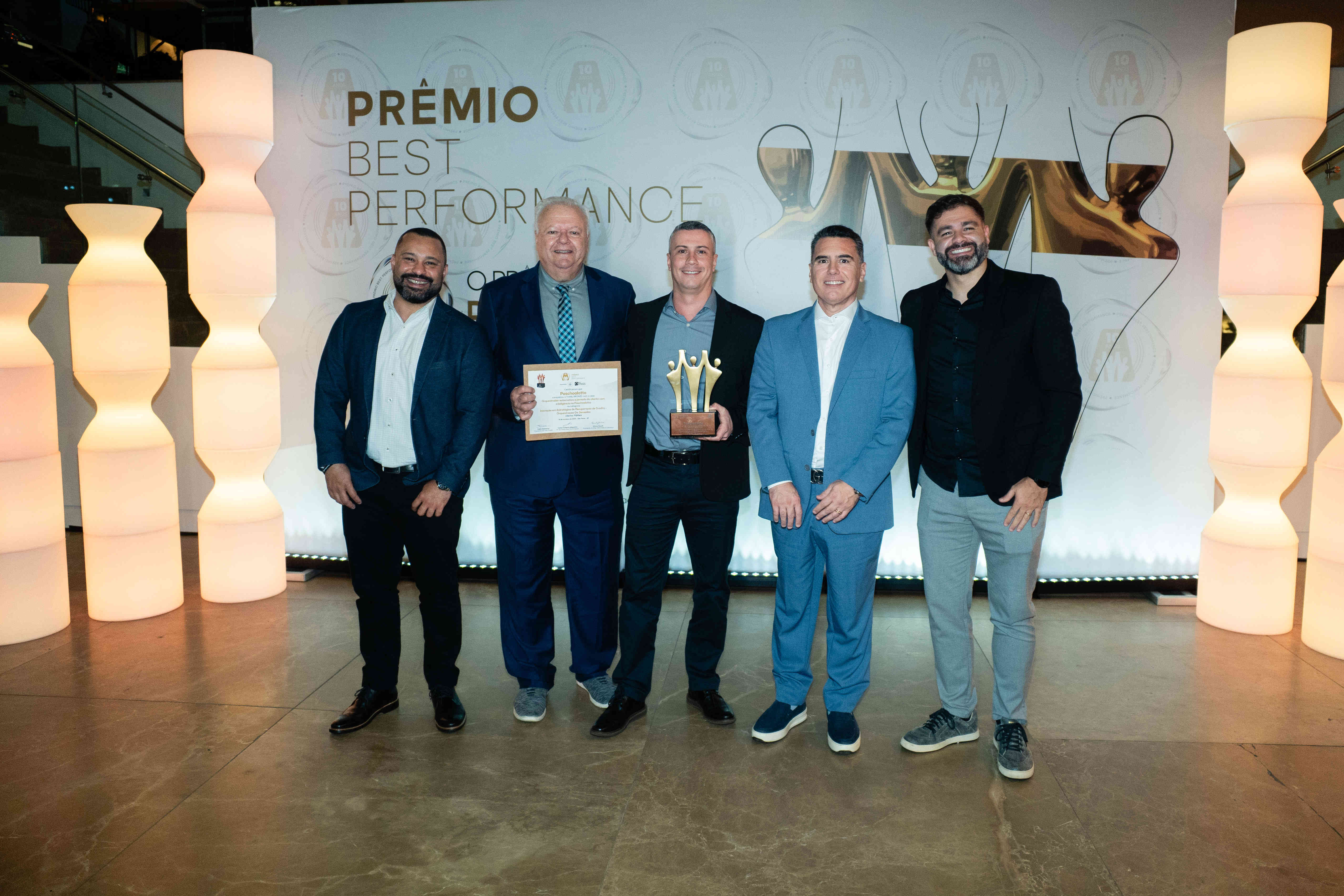 Executivos da Paschoalotto posando com troféu bronze e certificado no palco do Prêmio Best Performance 2025