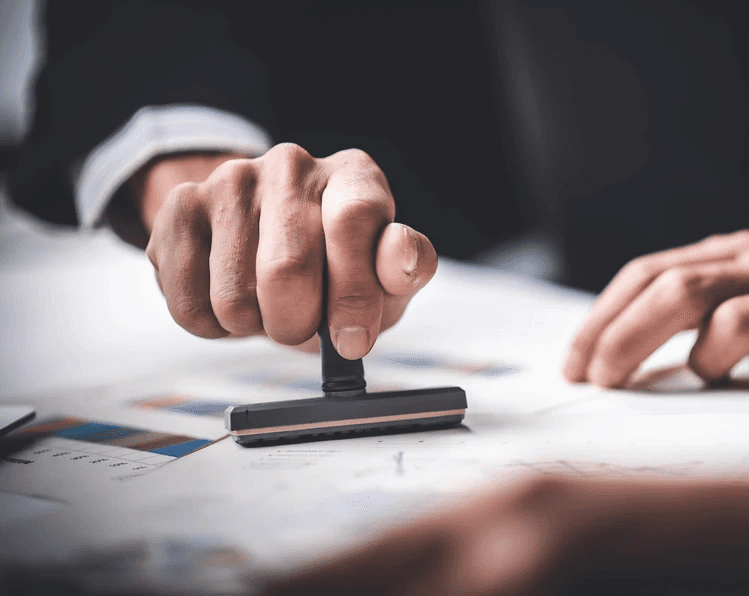 Mão segurando um carimbo contra um papel, como se aprovando um projeto