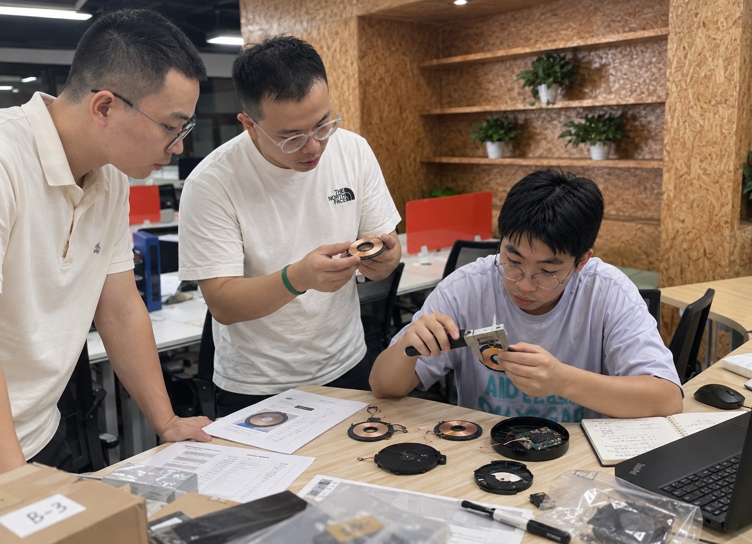Three people inspecting circular electronic components with a caliper at a worktable