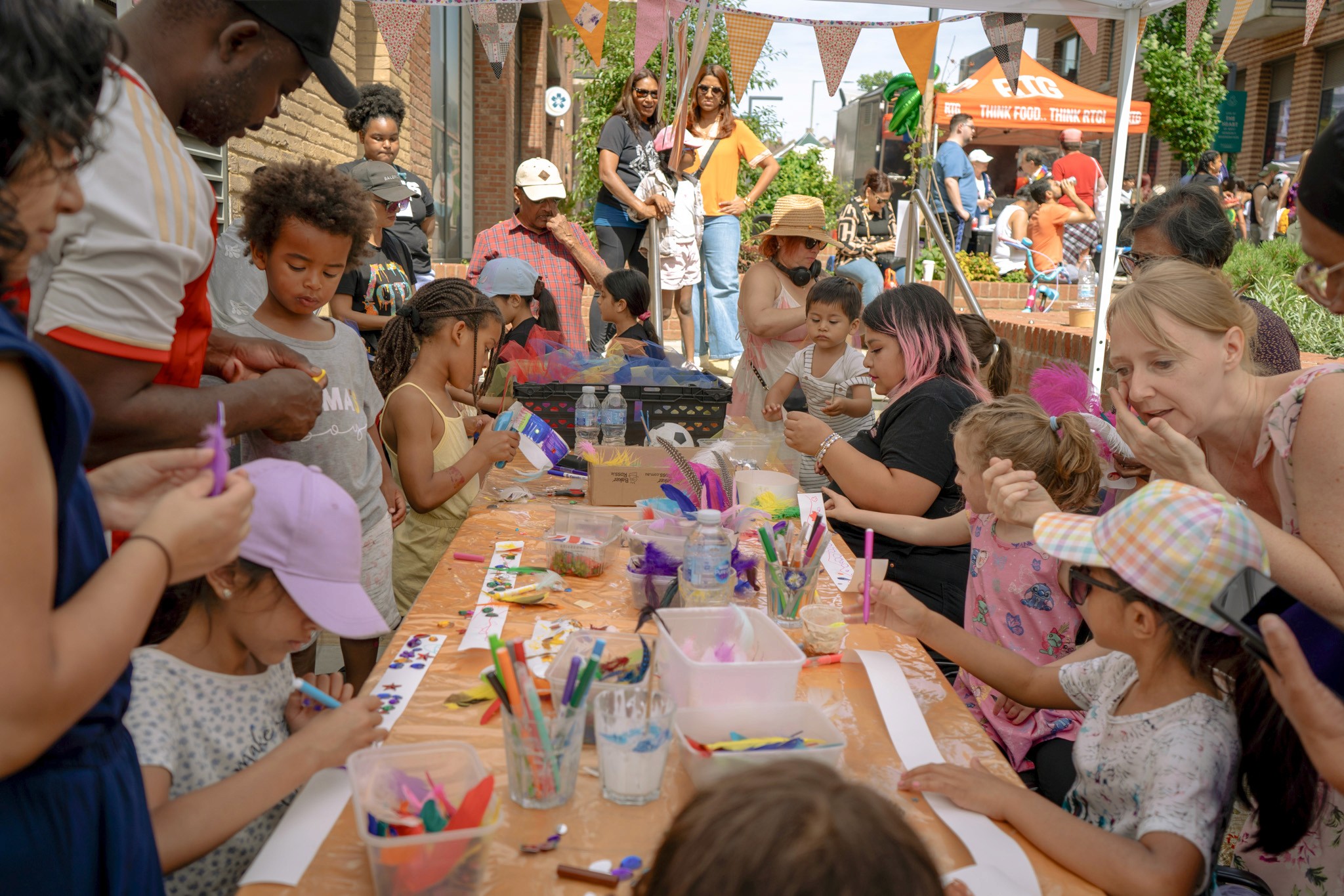 Children and adults sit and stand around a long craft table, drawing and decorating with colourful pens and feathers at an outdoor community carnival.