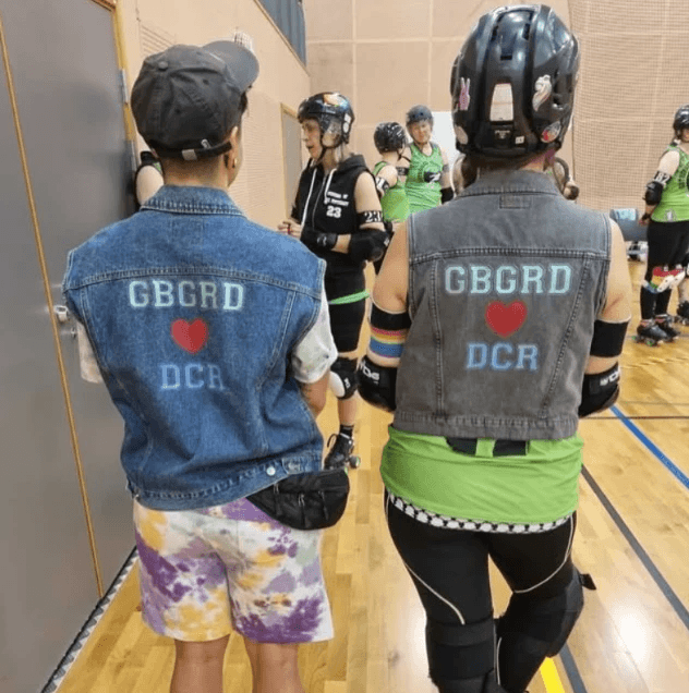 Two people standing with their backs turned towards the camera, with two jeans jackets saying GBGRD heart DCR.