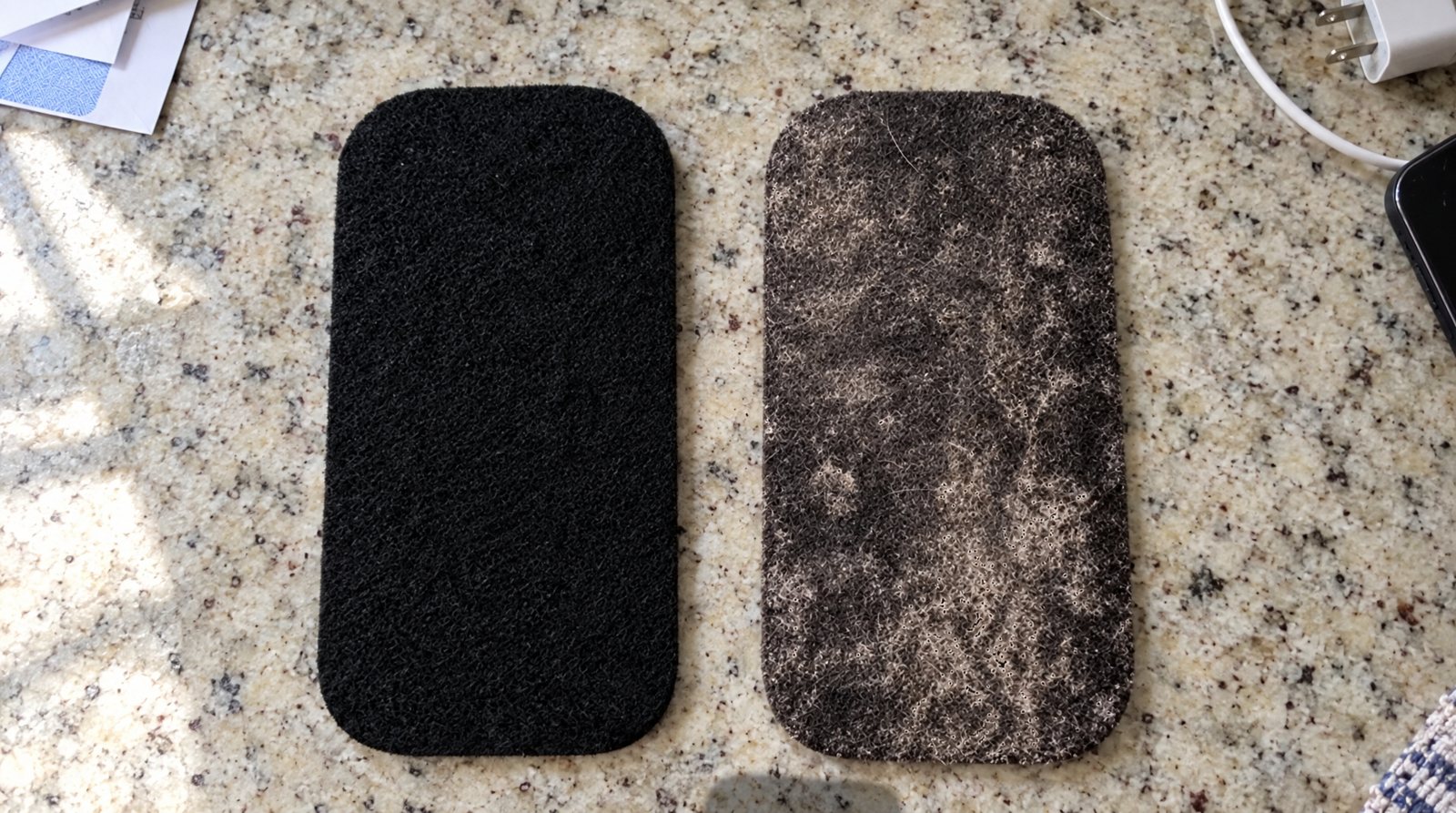 A new black Barnakl Pearl pad next to one after 30 days, coated in grey dust, on a kitchen counter.