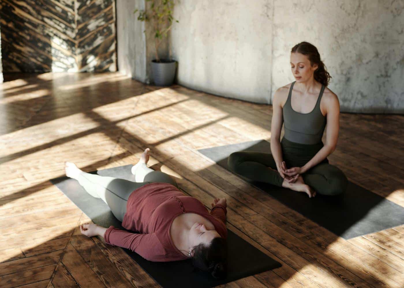 Woman laying down on mat with other woman sitting cross-legged on another mat guiding her