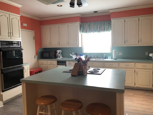 A spacious kitchen features pastel-colored walls, white cabinetry, and a large central island with wooden stools, set against a backdrop of a window with striped curtains allowing natural light to illuminate the room.
